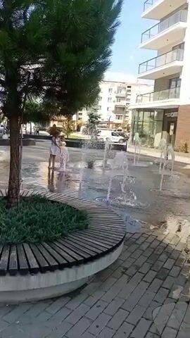 The video shows an outdoor urban scene with a paved plaza and a modern building in the background. In the foreground, a circular planter with green foliage is visible. Several children are playing in a water fountain feature that sprays water upwards from the ground. The children are dressed in summer clothing. The sun is shining, suggesting it is daytime. The environment appears to be a public space.