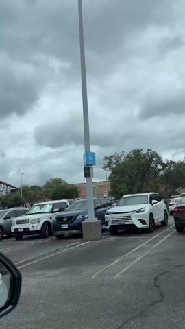 The camera, positioned from inside a car, pans across a parking lot on a cloudy day. Various cars are parked, including a white Land Rover, a blue Nissan Pathfinder, a red Toyota, and other assorted vehicles. The camera moves to the right, showing a covered entrance with architectural pillars and a wooden roof. A person is visible exiting one of the cars. A navigation system announces instructions in Spanish.