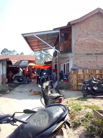 A street scene with several motorcycles, some parked and one in the foreground with a black and yellow seat. To the left, a market is set up with several stalls covered by orange tarps, and people are visible browsing. A brown dog lies on the ground in the middle of the street. To the right, a two-story brick building stands with a covered patio area. Wooden crates are stacked against the side of the building. The sky is clear and the sun is shining.