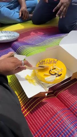 The video shows a group of people sitting on a colorful mat outdoors, likely for a picnic or celebration. A yellow cake with "MAYUR" written on it in yellow icing is visible in the foreground. One person is using a wooden utensil to cut a slice of the cake. In the background, two men are visible, one is holding a phone as if taking a photo or video, and the other appears to be clapping or gesturing. The environment suggests a natural setting with rocks and water in the distance, possibly a riverbank. The overall scene conveys a festive atmosphere with a birthday celebration.