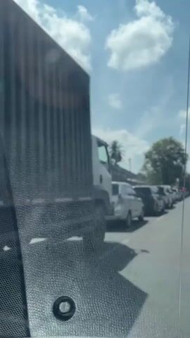 A line of cars and a truck are stopped on a paved road under a bright, cloudy sky. Palm trees and houses are visible on the left side of the road, while a large tree with full foliage is on the right. The camera is inside a vehicle, looking forward through the windshield. The vehicles are in a single lane, suggesting a traffic jam.