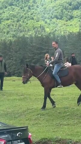 The video shows a man riding a horse on a grassy field with a forest in the background. The man is wearing a gray sweatshirt and jeans. The horse is brown and has a saddle. Another horse is also present and is being held by a man wearing a vest. There are three women standing nearby, with two of them using their phones, appearing to be taking pictures. A black car is parked in the foreground, and its license plate is partially visible. The environment appears to be a rural or mountainous area.