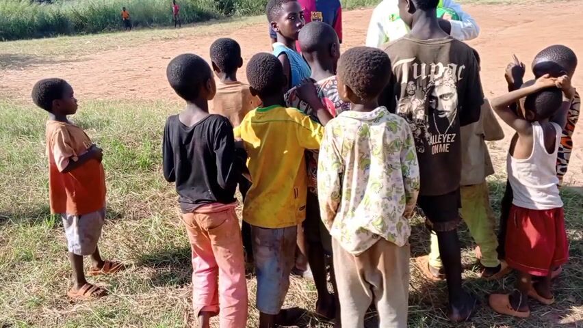 A group of children of varying ages are gathered in a small cluster, primarily facing a man in a white shirt and yellow safety vest. Another taller boy stands with him, wearing a soccer jersey. The man holds up a light blue soccer jersey with "HAALAND 9" printed on the back. The children are looking at the jersey with interest. The scene takes place on a dirt field with green grass and trees in the background. There are hills and mountains visible in the distance.