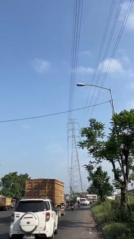 The video shows a street scene with a clear blue sky dotted with scattered clouds. A large power transmission tower stands prominently in the background, with several high-voltage power lines extending horizontally across the frame. A street light is attached to a pole on the right side of the road. Several vehicles are present on the asphalt road, including a white SUV in the foreground, a large dump truck with a brown cargo bed filled with sand, and other cars and motorcycles further down the road. Trees with green foliage are visible on the sides of the road and in the background. The overall impression is a busy road on a sunny day, possibly in an urban or semi-urban area, with infrastructure for electricity transmission.