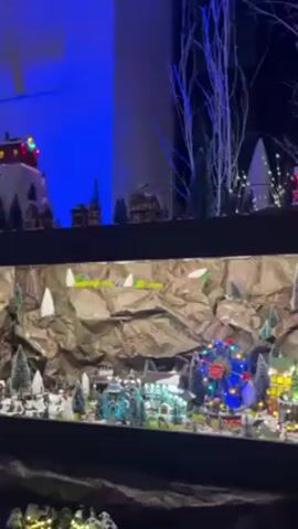 The video shows a multi-tiered Christmas village display. The top tier is lit with blue light and features buildings on a snowy hill, plus decorative white branches with small lights. A ski lift carries yellow cars across the display. The middle tier features a mountain backdrop made of crumpled brown paper, with artificial snow patches and trees. Below, a miniature village includes lit buildings, trees, and a Ferris wheel lit with colorful lights. The bottom tier is partially visible and seems to continue the snowy village theme. The entire display is enclosed in a dark cabinet or shelf.
