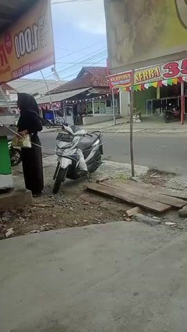 A motorbike is parked on the side of the road. A woman wearing a black hijab and dress is standing on the sidewalk. Two women in pink hijabs and light-colored dresses are walking across the street. There are shops in the background with signs that say "SERBA 35.000." The weather appears to be overcast.