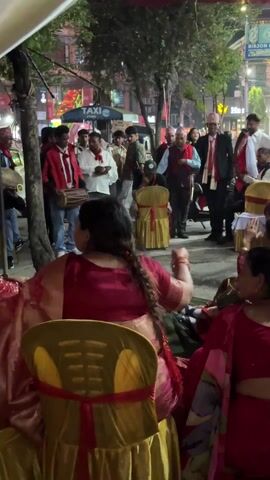 The video captures an evening outdoor gathering in an urban street of Pokhara, Nepal. Numerous people are present, including men wearing suits often paired with traditional Nepali Dhaka topi-like hats, and many women seated on the ground dressed in red and maroon traditional attire. Some men are seen with drums, indicating musical performance or celebration. A woman in a white jacket, carrying a baby, walks towards a man and briefly embraces him. The scene is lit by artificial lights, and background elements include street signs for "TAXI," "TUBORG," and "BIRJON FAST FOOD." The overall atmosphere appears social and festive.