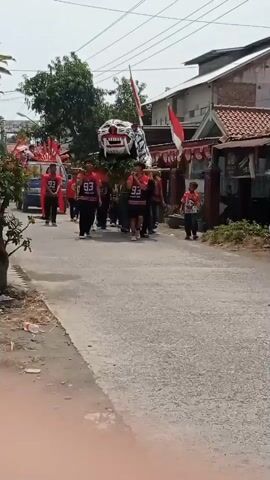 A group of people are walking down a street, carrying a large tiger effigy. The tiger effigy is white with red stripes and has a fierce expression on its face. The people carrying the effigy are wearing red shirts with the number 93 on them. There are also some Indonesian flags visible in the background. A few vehicles are also present on the street, including a motorcycle and a car. The street appears to be a residential area, with houses lining both sides of the road. The sky is clear and it appears to be a sunny day.