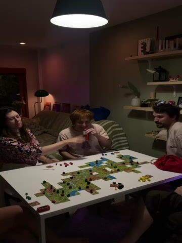 A group of four people are playing a board game at a white table in a dimly lit room. The game appears to be Carcassonne, with tiles laid out on the table forming a map of cities, roads, and fields. Two people are visible on the left side of the table, one woman with dark hair wearing a colorful floral shirt, and another person with reddish-blond hair wearing a pink t-shirt, holding a red cup. On the right side of the table, a man with dark hair and a beard is leaning over the game. Another person's legs are visible in the foreground on the right. The room has a couch in the background with colorful pillows, a floor lamp, and shelves with various items on the wall. The lighting is low, with a large pendant light hanging overhead casting a bright glow on the table. The overall atmosphere suggests a casual gathering of friends playing a game.