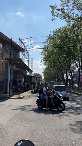 A motorcycle with two people on it rides down a paved road. The person in the front is driving and wearing a helmet. The person behind is wearing a white hijab. They are both wearing casual clothes. The road is lined with trees on the right and buildings on the left. The buildings appear to be shops. There are power lines overhead. A silver car is parked on the side of the road behind the motorcycle. The sky is blue with some clouds. The overall impression is a sunny day in a somewhat urban or suburban environment.