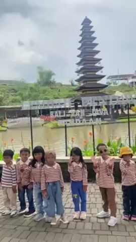 The video shows a group of seven young children posing for a photo in front of a lake. The children are all wearing matching striped shirts. Behind the children is a lake with a tiered pagoda structure on an island in the middle of the lake. There is a sign in front of the lake, and vegetation around it. The sky is overcast. The children are all smiling and waving at the camera.