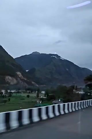 The video shows a view from inside a vehicle, looking out at a mountainous landscape under a cloudy sky. A road with a white and black striped barrier runs alongside the vehicle. Green fields and scattered buildings are visible in the valley. A large, tree-covered mountain dominates the background. Several vehicles, including a truck and a white car, are seen on the road. Reflections from the vehicle's interior are visible on the windows.
