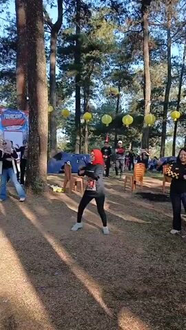 The video shows an outdoor gathering in a wooded area with tall trees and dry grass. Yellow lanterns are strung between the trees. In the foreground, two women are dancing. The woman on the left wears a red headscarf, a black long-sleeved shirt, black pants, and white sneakers. The woman on the right wears a black and yellow checkered long-sleeved shirt, blue jeans, and white sneakers. In the background, several tents are set up, and a group of people are gathered. Some people are standing, while others are sitting in chairs. There are also several orange plastic chairs scattered around the area. The ground is covered in dry leaves and pine needles. The sun is shining, casting shadows through the trees. The overall atmosphere appears to be festive and casual.