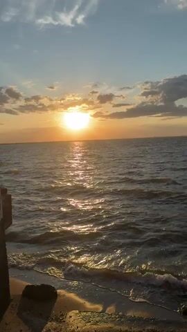 The video captures a tranquil sunset over a body of water. The sun, a bright orb, is partially obscured by scattered clouds, casting a warm, golden glow across the rippling surface of the water. The light reflects off the waves, creating a shimmering effect. On the left side of the frame, a small section of a sandy shore is visible, with some rocks and what appears to be a concrete structure. The sky transitions from a bright blue near the top to softer hues of orange and yellow near the horizon, a typical characteristic of a sunset. The overall atmosphere is peaceful and serene.