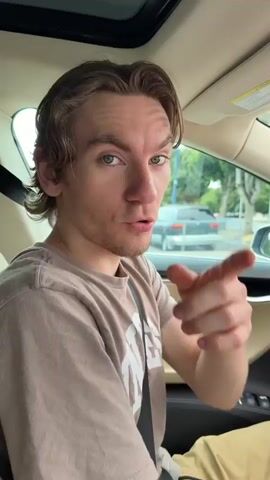 A young man with blue eyes and brown hair is sitting in the driver's seat of a car. He is wearing a tan t-shirt and tan pants. The man is speaking directly to the camera, gesturing with his hands. In the background, there are cars and trees visible through the car windows. The man appears to be making a threat or demand.