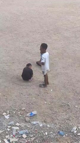 The video shows three children playing on a dirt patch next to a pile of trash. One child, wearing a white shirt and shorts, is standing and holding a stick, which he is pointing at another child who is squatting on the ground. The third child is sitting on the ground and holding a toy gun. The ground is covered with dirt, small rocks, and trash.