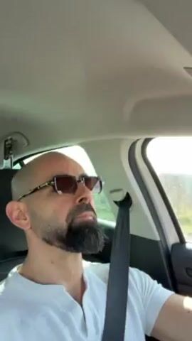 A man with a shaved head, beard, and sunglasses drives a car while listening to music. He is wearing a white shirt and a seatbelt. The scenery outside the car appears to be rural. He occasionally points at the ceiling of the car.