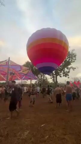 The video shows a festival with music, dancing, and a large, stationary, striped hot air balloon. There are many people present, some of whom are shirtless. The ground is brown and possibly muddy. There's a colorful fabric structure on the left side of the frame. The sky is partially cloudy.
