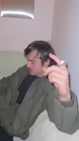 The video shows a man lying on a light-colored sofa, wearing a black t-shirt and an olive green jacket. He appears to be either sleeping or resting with his eyes closed. The camera is unsteady and pans around the room, revealing a wall with a brown rectangular fixture and other objects out of focus in the background.