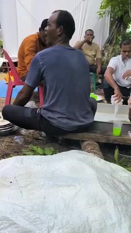 A group of men are gathered outdoors, possibly for a communal activity or discussion. One man in a white shirt is actively engaged with a white container, appearing to be washing or cleaning something with a white cloth. Another man, wearing a light brown shirt, is seated and observing the activity. A third man, in a blue shirt, is visible in the background, looking towards the central action. In the foreground, a white tarp or large bag is spread out, and some green leaves are scattered on the ground. The environment appears to be somewhat rustic, with trees and a rudimentary structure suggested in the background. There are also some cardboard boxes and a green cup on a wooden plank. The overall scene suggests a casual, outdoor gathering.