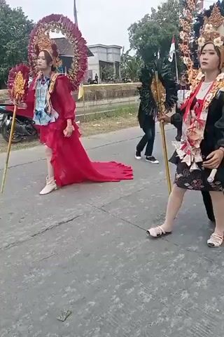Two individuals are participating in a parade, dressed in elaborate costumes. The person on the left wears a vibrant red dress with a long train and an ornate, fan-like headdress adorned with feathers. They are holding a long, slender golden staff. The person on the right is dressed in a black outfit, also featuring a large, circular headdress embellished with gold and black feathers, and a dramatic train. They too are holding a golden staff. Both individuals appear to be posing for the camera, with spectators and a few motorcycles visible in the background on a paved road. The environment suggests an outdoor event in a somewhat rural or suburban setting, with a cloudy sky overhead.
