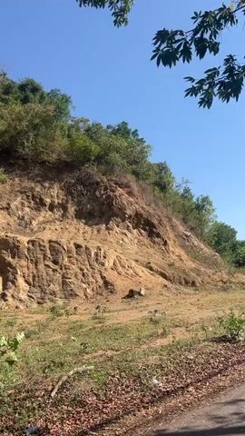 This is a handheld, low-angle outdoor video showing a steep, rocky hillside with vegetation on top and in the foreground. The sky is blue with some scattered clouds. The camera pans slightly to the right. There are no people or animals visible in the video. The environment appears to be a rural or natural setting, possibly a roadside cut.