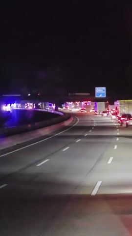 This video shows a highway at night. Emergency lights, blue and red, are flashing on the left side of the road, illuminating multiple vehicles and what appear to be emergency personnel. Traffic is backed up, with numerous cars and trucks stopped in the lanes. The sky is dark, and the road is illuminated by the emergency lights and vehicle headlights. A highway sign is visible in the distance.