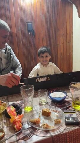 A young boy with short, dark hair and a white and yellow hoodie sits at a black Yamaha keyboard, smiling. To his left, an older man with gray hair, wearing a gray jacket, holds a lit cigarette. They are at a table covered with a patterned tablecloth, on which there are various glasses with yellow liquid, a bowl of sugar, a plate with remnants of food, and a pack of cigarettes. The wall behind them is paneled with wood. The boy appears to be playing the keyboard, and background music is audible. The man is looking at the boy and seems to be enjoying the performance.