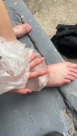 The video shows a person sitting outdoors with bare feet on a concrete surface, possibly a curb or low wall. The person is putting a clear plastic bag over their left foot. A black backpack is partially visible in the background. The scene appears mundane and straightforward.