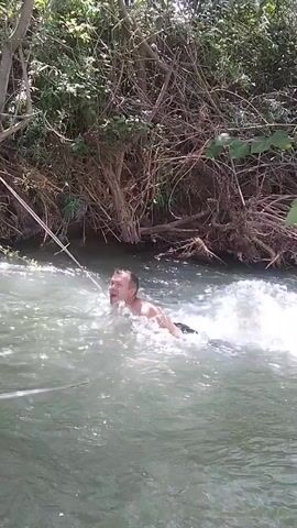 A man swims in a river. He is partially submerged in the water, with his upper body visible. He is looking to his right and appears to be struggling against the current. There is a rope visible to the left of the man, extending out of frame. The river is surrounded by lush green vegetation, including trees and bushes. The water is choppy and appears to be moving quickly. The sunlight is dappled, suggesting it is daytime.