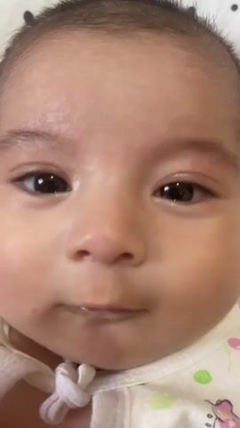 A close-up video shows a baby's face. The baby has dark eyes and a small mouth. The baby's mouth moves slightly as if making a sound or trying to speak. The baby is wearing a white onesie with a pattern of animal figures and dots. The background is out of focus and appears to be a soft, light color. The lighting is even and natural.