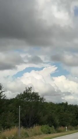 The video shows a view from a vehicle driving on a road, looking out at the sky and trees. The sky is filled with a mix of dark gray and white clouds, with patches of blue sky visible. There are trees and foliage lining the sides of the road. Several cars are also visible on the road ahead. The video appears to be shot on a cloudy day.