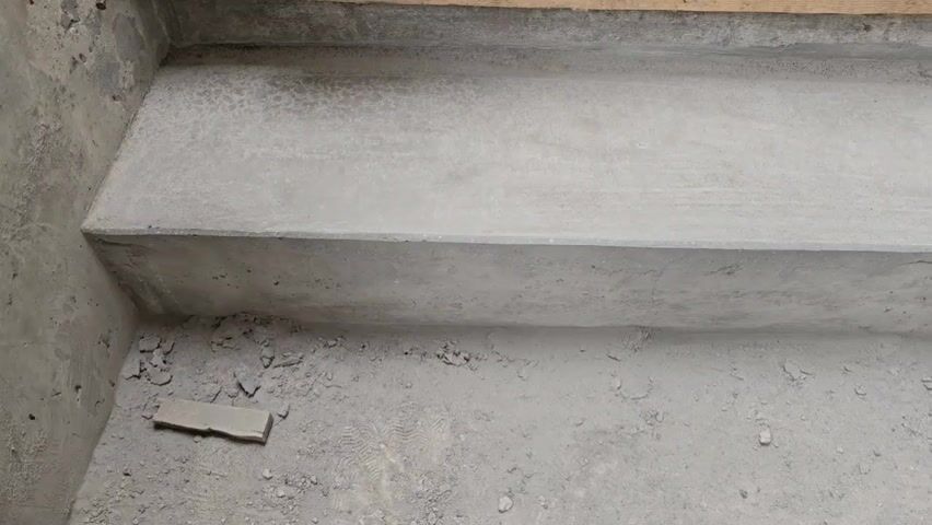 The camera focuses on a newly constructed gray concrete step, appearing rough and unpolished. Debris and a small flat piece of material, possibly concrete or tile, rest on the ground below the step. Above, a wooden plank indicates further construction is in progress. The lighting is ambient, casting soft shadows and highlights on the step's surface.
