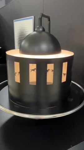 A black, cylindrical zoetrope with a black dome on top, mounted on a silver tray, is displayed on a black surface. Inside the cylinder, illuminated from above, are cutouts of horses in various stages of motion, creating an animation of running horses when viewed through the vertical slits in the cylinder. The zoetrope is in operation, showing the illusion of movement.