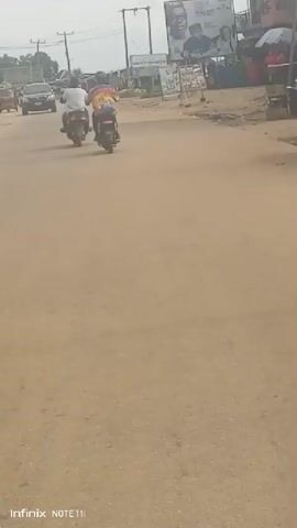 The video shows an outdoor street scene. Two people are riding motorcycles away from the camera. Other vehicles are visible, including a car and a "keke," a type of auto-rickshaw. There are buildings and utility poles on the sides of the street. A large advertisement billboard is also visible on the right side of the street. The street is paved, but appears slightly sandy or dusty. The sky is overcast. The footage appears to be shot from within a vehicle. The lower left corner shows the imprint "Infinix NOTE 11i."