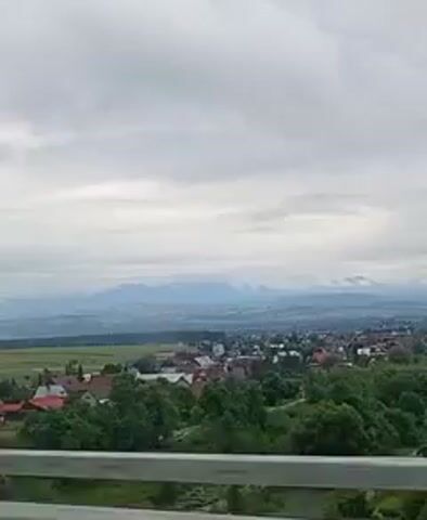 A high-angle medium shot shows a cloudy sky above a distant mountain range. Below the sky, a village with red-roofed houses is visible, surrounded by trees and fields. In the foreground, a metal railing partially obstructs the view. The camera is stationary.
