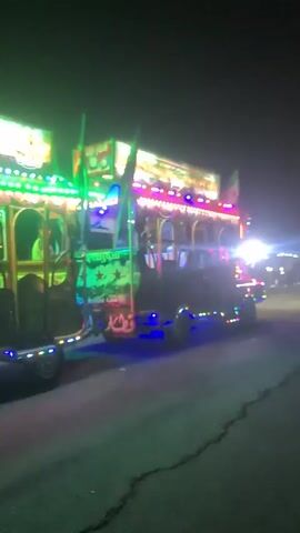 The video shows a bus decorated with bright neon lights driving down a dark road at night. The bus is multi-colored, switching between green, purple, and blue hues. It is followed by a trailer, also decorated with lights.  Other vehicles and some people can be seen indistinctly in the background, illuminated by the bus's lights. The road is asphalt and shows a crack running along the side.