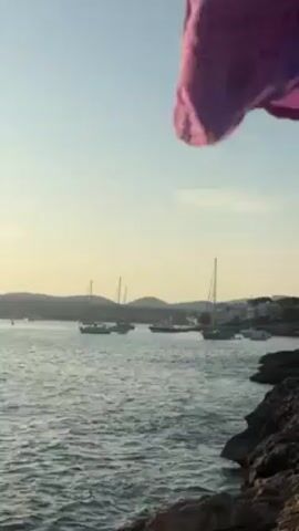 Okay, here is a description of the scene:

The video shows a coastal view, likely during sunset or early morning, as indicated by the soft, warm light. The foreground features a rocky shoreline. The sea, slightly choppy, extends towards the horizon, which is marked by low, green hills. Several sailboats are anchored in the water. The sky is mostly clear with a light blue hue. The overall atmosphere seems tranquil.