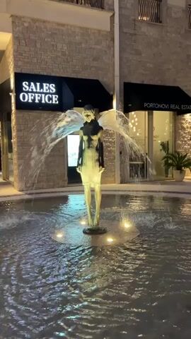 This video shows an outdoor fountain at night. The central subject is a bronze sculpture of a winged female figure, appearing to be an angel. The sculpture is illuminated by lights within the fountain's basin and also by external lighting, giving it a striking appearance amidst the flowing water. The water jets out from behind the sculpture, creating a dynamic visual effect that mimics wings or a flowing cloak.

Behind the fountain, there are buildings with stone facades and dark awnings. One awning has prominent white text that reads "SALES OFFICE". Another awning has text that is partially visible, appearing to say "PORTONOVI REAL ESTATE". These signs suggest the location is a commercial area, possibly a sales center for a real estate development. The overall ambiance is elegant and somewhat dramatic due to the lighting and the nocturnal setting. The water in the fountain is slightly rippled, reflecting the lights and the sculpture. The time of day is clearly night, and the weather appears calm as the water is not being significantly disturbed by wind. The scene evokes a sense of artistic beauty and perhaps a touch of mystery.