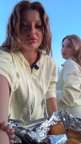 A woman is standing outside in a light yellow hoodie, holding a container of yellow sauce and a tray of food covered in foil. She dips her finger in the sauce, tastes it, and smacks her lips. Behind her, another woman in a matching outfit walks away towards a body of water. The background shows grass, a body of water, and a blue sky.