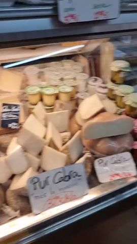 The video shows a display of cheeses and cured meats in a refrigerated case, likely at a market or deli in Setcases, Spain. The camera pans across the various products, highlighting an assortment of cheeses in different shapes, sizes, and textures, some with visible blue veins, and others appearing firm and aged. Several small tubs of what seem to be yogurt or custard are also visible.

Interspersed with the cheese are various types of cured meats, including sausages in rings and sliced charcuterie. The products are arranged neatly within the display case, with some items labeled. Visible text on the labels includes "Per Cornetto", "Ovella", "Ovella i Vaca", "Gourmet", "Bull Blanc", "Bull Negre", and "el mella". The lighting suggests an indoor setting, and there are no clear indicators of the time of day or weather. The overall impression is a well-stocked and appealing selection of artisanal food items.
