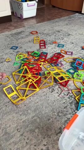 A young person is sitting on the floor, playing with colorful magnetic building tiles. The tiles are scattered across a gray carpet. There are many square, triangle, and hexagonal shaped tiles in red, yellow, green, blue, and purple. The person is holding some of the tiles and appears to be in the process of building something, or taking it apart. In the background, there is a white toy bin with toys inside, a cabinet, and a doorway leading to another room.