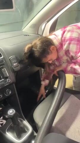 A young woman is vacuuming the interior of a car. She is wearing a pink and white plaid jacket, dark leggings, and has brown hair tied back in a ponytail. She is bent over, vacuuming the area around the driver's seat. The car is a white Volkswagen Polo with a manual transmission. The interior is black and gray. The car is parked in a garage or car wash.