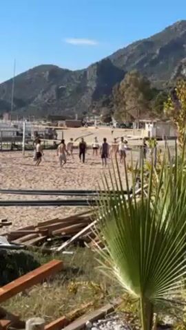 A group of young men are playing volleyball on a beach. They are spread out across the sandy court, interacting with each other as they volley the ball over the net. The backdrop consists of lush green mountains, a clear blue sky, and palm trees, which partially obscure the view from the foreground. Some wooden construction materials are scattered at the bottom of the screen.