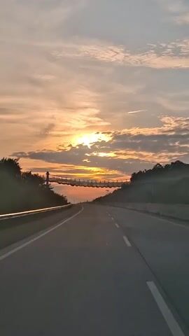 The video shows a view from inside a vehicle traveling on a highway. The sun is setting, casting an orange and yellow glow across the sky, with clouds scattered throughout. A pedestrian bridge is visible spanning the highway in the distance. Trees and foliage line the sides of the road. The road is paved with white lane markings.