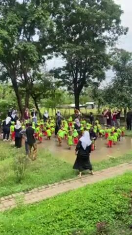 The video shows a group of children in red shorts and lime-green shirts standing and playing in a muddy puddle. Around them are adults, likely teachers or chaperones, some taking pictures. The area is surrounded by grass, trees, and a brick pathway. There is singing and music playing in the background. The setting seems to be an outdoor park or a garden-like area.