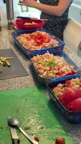 The video shows a kitchen scene where someone is preparing food. The focus is on three blue glass dishes filled with a seafood mixture containing shrimp, imitation crab meat, grapes, and other ingredients. Tomatoes are added as a garnish to one dish, while green onions top another. A person wearing a floral print dress is partially visible in the background, holding a container, potentially adding ingredients to the mix. A green cutting board and several utensils are visible on the counter. The environment is a well-lit kitchen with granite countertops.