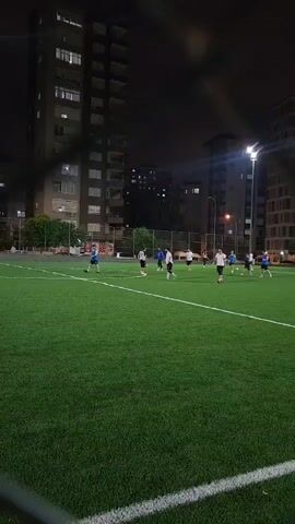 The video shows a nighttime view of a soccer field with artificial turf. Several people, likely players, are scattered across the field, engaged in a game or practice. They are wearing athletic attire, with some in blue shirts and others in white or black shirts. The field is illuminated by bright floodlights, casting long shadows and highlighting the green turf. In the background, tall residential buildings stand against the dark night sky, with some windows showing lights. A fence is visible in the mid-ground, separating the field from the surrounding area. The camera appears to be positioned behind a blurred foreground element, possibly a fence or netting, which frames the scene. The overall atmosphere is one of casual athletic activity taking place in an urban, nighttime setting.