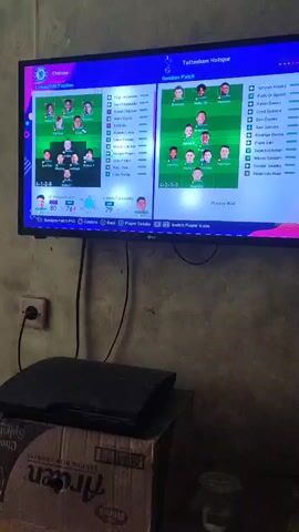 The video shows a television screen displaying a soccer video game. The game appears to be a simulation or management game where players can set up their teams and strategies. The screen is split into two halves, with the left side showing the Chelsea team lineup and the right side showing the Tottenham Hotspur team lineup. Both teams are displayed in a 4-1-2-3 formation with player portraits and names visible. Below the team lineups, there are game interface elements, including buttons and text labels that suggest options for managing players, changing formations, and starting the match. The television is mounted on a wall, and below it, a black gaming console, possibly a PlayStation 3, sits on top of a cardboard box. The overall environment appears to be a room with a plain wall, suggesting a domestic setting. The video is shot from a slightly low angle, looking up at the television.
