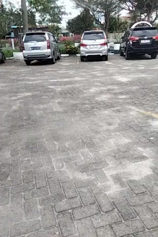A security camera captures an outdoor view of a parking lot. Several cars are parked in a row, some are silver and one is dark colored. The cars are parked on a paved surface made of rectangular stones. Trees and some vegetation are visible in the background. The sky appears overcast. The camera angle is tilted, giving a vertical perspective of the scene.